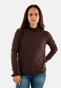 Maglione marrone lavorato a maglia con pois, colletto alto a ruches, maniche lunghe e polsini arricciati. Presenta piccoli bottoni all'orlo. Abbinato a jeans blu.
