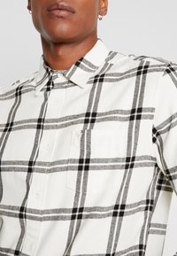 Gros plan d'une personne portant une chemise blanche à motif à carreaux noirs et gris avec une poche poitrine, et de petites boucles d'oreilles en anneau visibles.