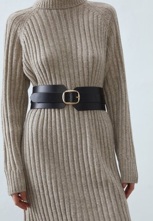 Beżowa sukienka dzianinowa z prążkowaną fakturą i golfem, przewiązana w talii szerokim czarnym paskiem z złotą kwadratową klamrą.