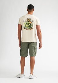 Cremweißes Baumwoll-T-Shirt mit buntem Kaktus-Design und dem Schriftzug "BAJA CALIFORNIA." Kombiniert mit olivgrünen Cargo-Shorts und weißen Sneakern.