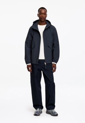 Homme debout portant une veste à capuche bleu marine, une chemise blanche à manches longues, un pantalon bleu marine et des chaussures de sport grises sur fond blanc.