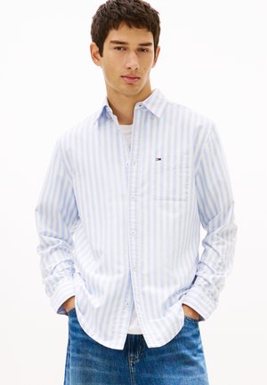 Ung mand iført en lyseblå og hvidstribet skjorte med knapper over en hvid T-shirt, med hænderne i lommerne og blå jeans.