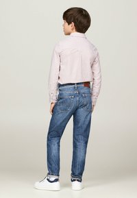 Ragazzo che indossa una camicia a righe con bottoni e jeans denim blu, abbinati a scarpe da ginnastica bianche. La camicia ha maniche lunghe e un dettaglio sullo yoke posteriore.