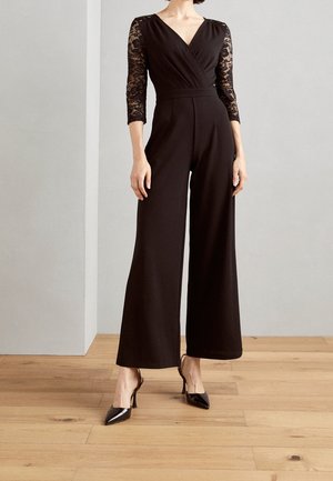 Vrouw in een zwarte jumpsuit met kanten driekwart mouwen, een V-hals, wijde pijpen en zwarte hoge hakken, staand op een houten vloer.