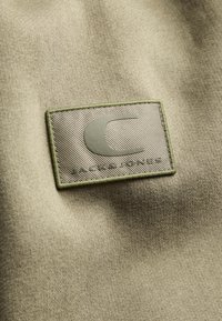 Beige fabric label with "C" logo and "JACK & JONES" text; bordered by green stitching; smooth texture with diagonal pattern on the label.