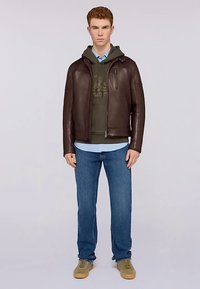 Brun läderjacka med dragkedja, över en grön huvtröja. Ljusblå skjorta, blå jeans och beige sneakers kompletterar outfiten.