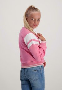 Jonge meid met een blonde paardenstaart, gekleed in een roze en witte jas en blauwe spijkerbroek, staande met een lichte glimlach en haar hand bij de kraag.