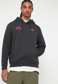 Tmavohnedá mikina s kapucňou so predným vreckom, šnúrkou na kapucni a červeným logom "ADIDAS ORIGINALS" na hrudi s trojdielnym akcentom nad ním.