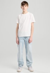 Valkoinen puuvillainen T-paita yhdistettynä vaaleansiniin farkkuehtoihin. Farkut ovat rento istuvuus ja niissä on haalistunut pinta. Valkoiset lenkkarit täydentävät asukokonaisuutta.