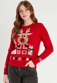 Pull en tricot rouge avec un motif de renne, des cornes pailletées, un nez en pompon et des motifs de cadeaux. Manches et ourlet côtelés. Jeans gris.