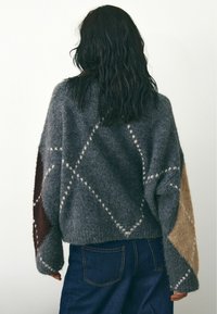 Pinto pilkas megztinis su laisvu siluetu, puoštas geometriniais raštais šviesiai pilkos, rudos ir smėlinės spalvų. Tekstūruota medžiaga ir per dideli rankovės.