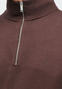 Brun strikket genser med høy krage, med en sølvglidelås og en liten brodert logo på venstre side. Myk tekstur.