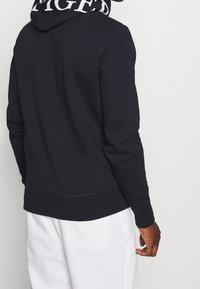 Sudadera negra con cordón y logotipo blanco en contraste en la capucha. La tela parece suave con una textura lisa. Silueta ajustada.