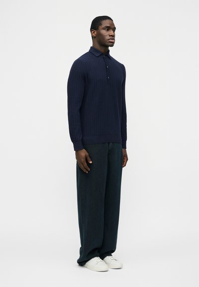 Homme debout portant un polo à manches longues côtelé bleu marine, un pantalon ample foncé et des baskets blanches, sur un fond clair uni.