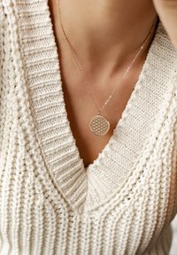 Delikatny srebrny naszyjnik z okrągłym wisiorkiem o geometrycznym wzorze, noszony na tle fakturowanego białego swetra z dekoltem w serek.