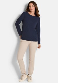 Frau trägt ein navyblaues Langarmoberteil, beige Hosen und hellbeige Sneaker, steht mit einer Hand hinter ihrem Rücken vor einem weißen Hintergrund.