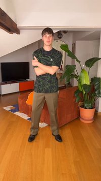 Ung mann iført trykt svart skjorte og brune bukser poserer med armene i kryss i en stue med oransje sofa og stor potteplante.