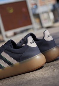 Par de zapatillas Adidas azules con rayas blancas y verdes y suelas marrones translúcidas sobre una superficie de hormigón al aire libre.