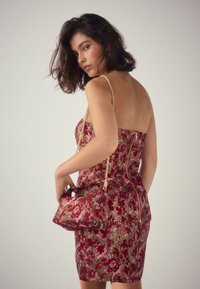 Frau posiert in einem trägerlosen roten und rosa Blumenkleid, hält eine passende Handtasche mit goldenen Kettenriemen vor einem schlichten Hintergrund.