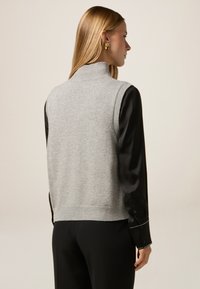 Suéter sin mangas de lana gris con un cuello alto, dobladillo acanalado y aberturas laterales, usado sobre una camisa negra de satén de manga larga.