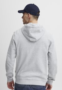 Sweat-shirt gris avec capuche, poignets côtelés et taille côtelée. Porté avec une casquette marine. Le tissu semble doux et texturé, adapté au port décontracté.