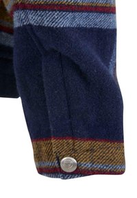 Ullskjorte med blå, rød og gul stripet mønster; har en sølvknapp ved mansjetten og en teksturert stofffinish.