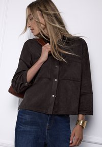 Camisa de ante marrón con botones, dos bolsillos en el pecho, mangas tres cuartos y herrajes plateados, combinada con vaqueros de mezclilla azul oscuro.