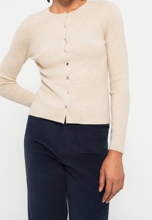 Kvinde iført en beige knappet cardigan og marineblå kordfløjlsbukser, stående med den ene hånd på hoften og den anden afslappet.