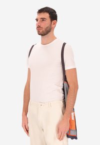 Uomo che indossa una t-shirt bianca e pantaloni crema, con una borsa a tracolla grigia con fodera arancione e manici scuri sulle spalle.