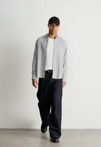 Gai pelēku svītru krekli, kas uzvilkts virs balta krekla, kopā ar tumšiem džinsu biksēm un melniem apaviem. Modelis valkā melnu cepuri.