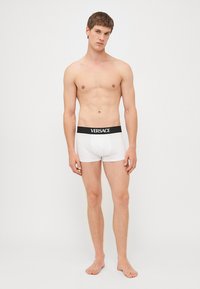 Weiße Boxer-Briefs mit einem schwarzen Versace-Logo-Bund, enganliegendem Design, glatter Textur und kurzem Bein. Model steht barfuß, Arme entspannt.