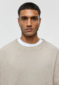 Beige gebreide trui met een losse pasvorm en ribbelstructuur, gedragen over een witte ronde hals t-shirt. Voorzien van een uitgesproken halslijn en oorbel.