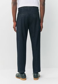 Dark blue denim trousers with a straight leg, featuring back pockets and a waistband. Worn with dark shoes and visible ankle.