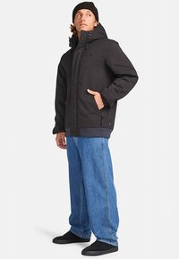 Vyras, dėvintis juodą kepurę, juodą striukę, plačius mėlynus džinsus ir juodus batus, stovi su viena ranka kišenėje ir žiūri į šoną.