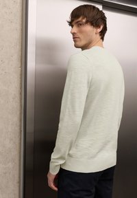 Jeune homme aux cheveux bruns courts portant un pull beige clair et un pantalon foncé, debout devant une porte d'ascenseur métallique.