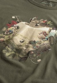 T-shirt verde oliva con un'illustrazione dettagliata di una scena interna accogliente con piante, mobili e due persone impegnate in attività.