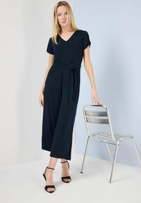 Navyblauer Jumpsuit mit kurzen Ärmeln, V-Ausschnitt, Taillenknoten und weit geschnittenen Beinen. Weicher Stoff mit einer glatten Oberfläche. Modell trägt schwarze Riemchenpumpen.