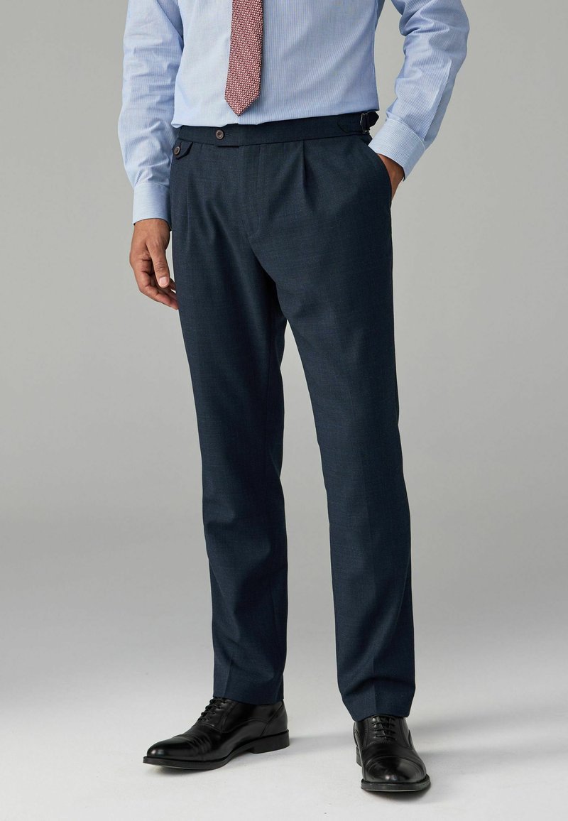 Pantalones ajustados de color azul marino con pliegues, fabricados en una tela texturizada. Presentan bolsillos laterales y adornos de botones. Combinados con zapatos de vestir negros.