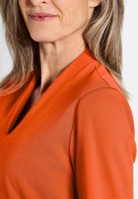Oranje, v-hals top van soepele, lichte stof. Heeft een gestructureerde schoudernaad en een nauwsluitende pasvorm rond de hals en schouders.