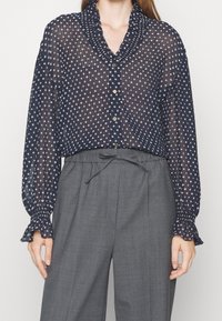 Blouse marine transparente à pois blancs, avec col froncé et fermeture à boutons, associée à un pantalon gris taille haute et manches froncées.