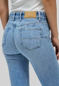 Jeans de mezclilla azul claro con un diseño ajustado, que cuentan con dos bolsillos traseros y una etiqueta de marca de cuero marrón en la cintura.