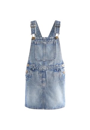 Ung pige smiler med flettet hår iført en lyserød langærmet top og blå denim overalls mod en neutral baggrund.