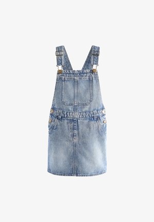 Ung pige smiler med flettet hår iført en lyserød langærmet top og blå denim overalls mod en neutral baggrund.