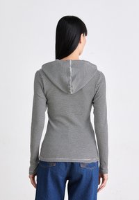 Svart og hvit stripete langermet hettegenser med en tettsittende design og teksturert stoff. Har hette og en liten utvidelse i bunnen.