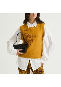 Gilet en maille duveteuse couleur moutarde avec broderies florales, associé à une chemise blanche, un pantalon à motifs et un petit sac à main noir.