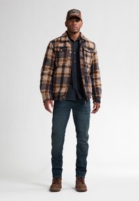 Rutet flanneljakke i brune og beige nuancer, mørk denimskjorte under, blå jeans, brune støvler og en beige kasket med logo detaljer.