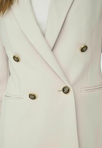 Blazer doppiopetto beige chiaro realizzato in tessuto morbido, con bottoni dorati con dettagli neri e tasche laterali.