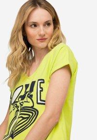 Camiseta de algodón verde lima con un corte relajado. Presenta un estampado gráfico negro de la palabra "AMOR" y elementos de diseño abstracto. Mangas cortas.