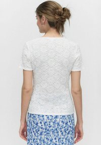 Hvid blondetop med korte ærmer, der har et struktureret blomsterdesign, parret med en blå nederdel med blomsterprint, set fra ryggen.