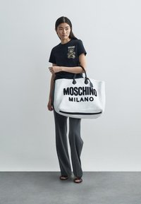 Λευκή τσάντα tote με μαύρο κείμενο "MOSCHINO MILANO", κατασκευασμένη από υφασμένα υλικά, με γκρίζες λεπτομέρειες και διπλές χειρολαβές. Το μοντέλο φοράει μαύρο t-shirt και παντελόνι.